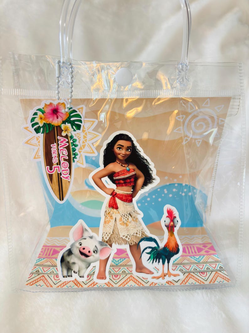 May include: A clear plastic tote bag featuring a Moana-themed insert with Moana, Pua the pig, and Heihei the rooster. A surfboard-shaped tag attached by a silver chain reads Melody Turns 5, decorated with a hibiscus flower and tropical leaves.