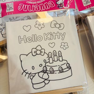 May include: A white canvas featuring a Hello Kitty design, including a cake and the text "Hello Kitty." The canvas is packaged in a clear plastic bag and includes a pink banner that says "Julianna" and "7th Birthday."