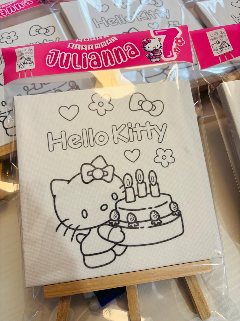 May include: A white canvas featuring a Hello Kitty design, including a cake and the text "Hello Kitty." The canvas is packaged in a clear plastic bag and includes a pink banner that says "Julianna" and "7th Birthday."