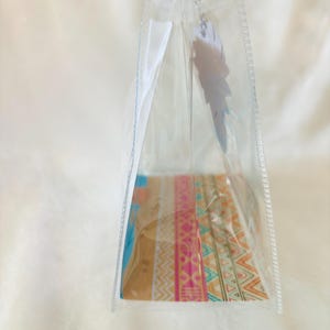 May include: A clear, triangular plastic pouch featuring a colorful base with horizontal bands of geometric patterns in shades of pink, orange, teal, and white. A white, feather-shaped tag hangs from the top zipper closure of the transparent bag.