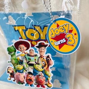May include: A clear plastic bag with a blue background featuring the word "TOY" in yellow. The bag has a tag that says "Joey turns 3". The bag is decorated with images of characters from the Toy Story movie.