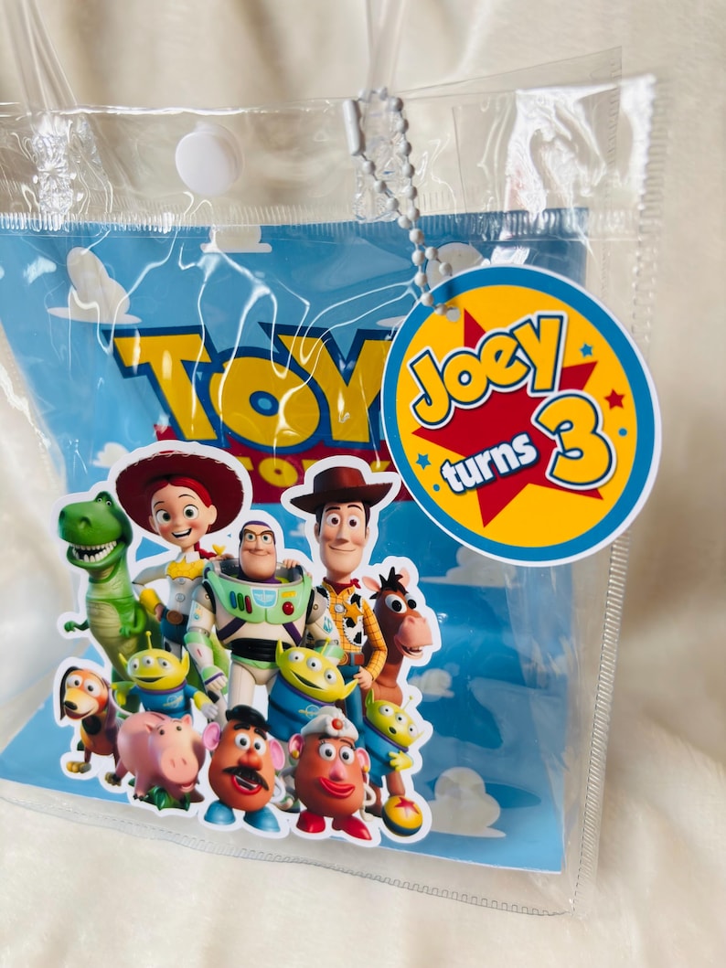 May include: A clear plastic bag with a blue background featuring the word "TOY" in yellow. The bag has a tag that says "Joey turns 3". The bag is decorated with images of characters from the Toy Story movie.