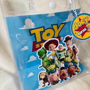 May include: Clear plastic gift bag featuring characters from the animated film Toy Story. The bag has a blue background with white clouds and the words "Toy Story" in yellow and red. A circular tag reads "Joey turns 3".