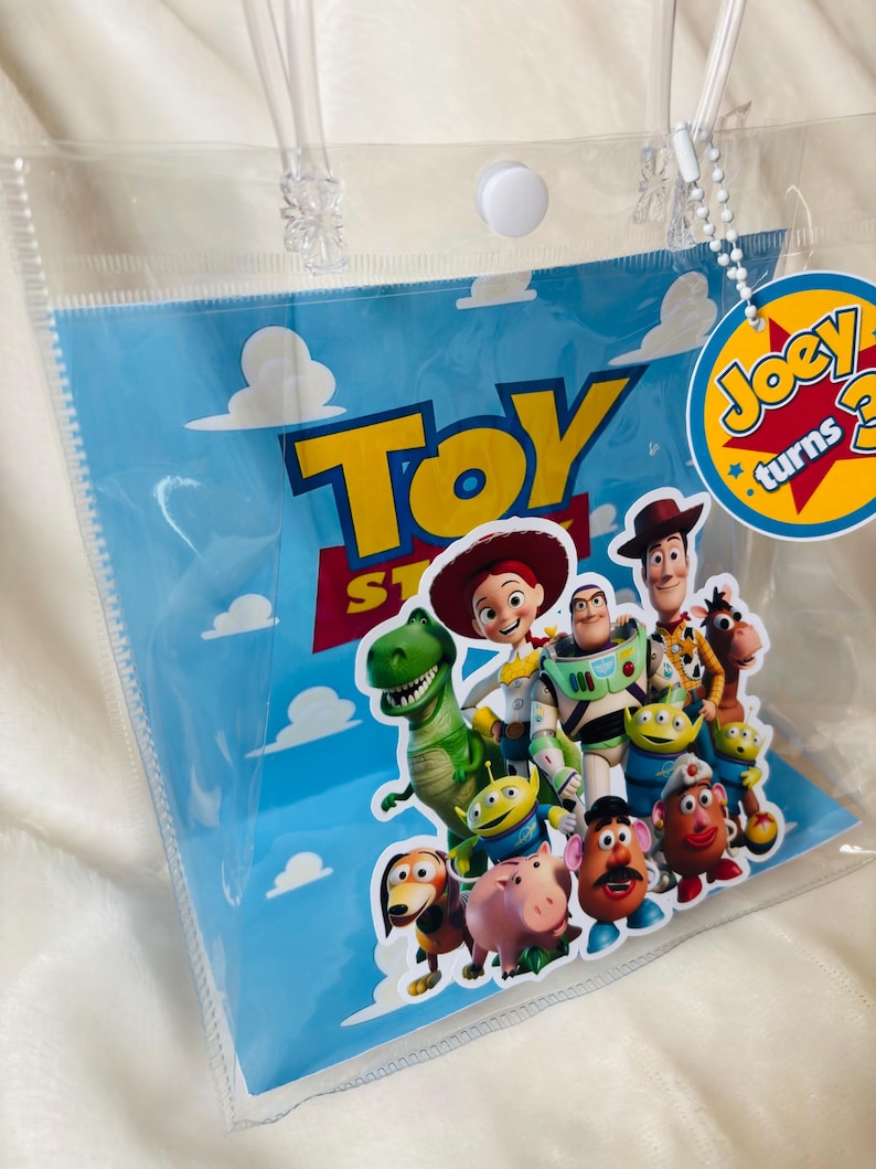 May include: Clear plastic gift bag featuring characters from the animated film Toy Story. The bag has a blue background with white clouds and the words "Toy Story" in yellow and red. A circular tag reads "Joey turns 3".
