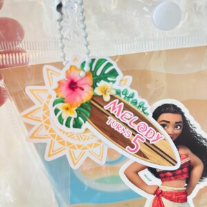 May include: A decorative tag featuring a surfboard graphic with the text Melody Turns 5, adorned with hibiscus flowers and tropical leaves, attached to a clear plastic bag by a silver ball chain, alongside a cutout of a long-haired character.