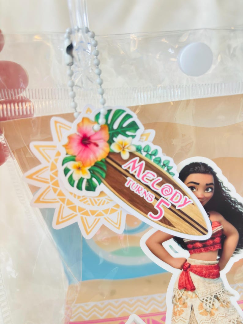 May include: A decorative tag featuring a surfboard graphic with the text Melody Turns 5, adorned with hibiscus flowers and tropical leaves, attached to a clear plastic bag by a silver ball chain, alongside a cutout of a long-haired character.
