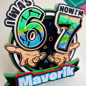 May include: A colorful birthday cake topper with the text "I WAS 6, NOW I'M 7" and the name "Maverik". The design features the numbers 6 and 7 in a graffiti style, with hands pointing towards the numbers. The color scheme includes green, blue, orange, and black.