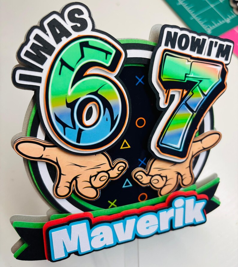 May include: A colorful birthday cake topper with the text "I WAS 6, NOW I'M 7" and the name "Maverik". The design features the numbers 6 and 7 in a graffiti style, with hands pointing towards the numbers. The color scheme includes green, blue, orange, and black.