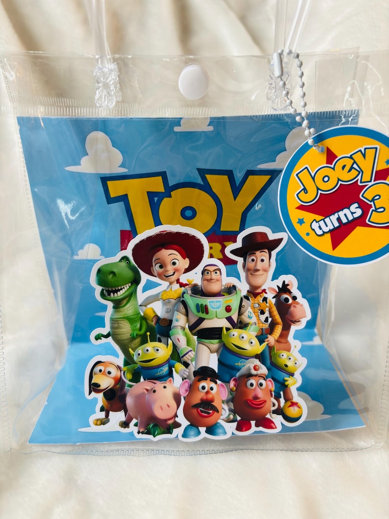 May include: A clear plastic bag featuring characters from the animated film "Toy Story." The bag has a blue background with the word "Toy" in yellow. A circular tag reads "Joey turns 3."