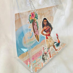 May include: A clear plastic tote bag with two transparent handles and a white snap closure. The bag features a decorative insert with Moana, Pua, and Heihei characters, plus a surfboard-shaped tag that reads Melody Turns 5.