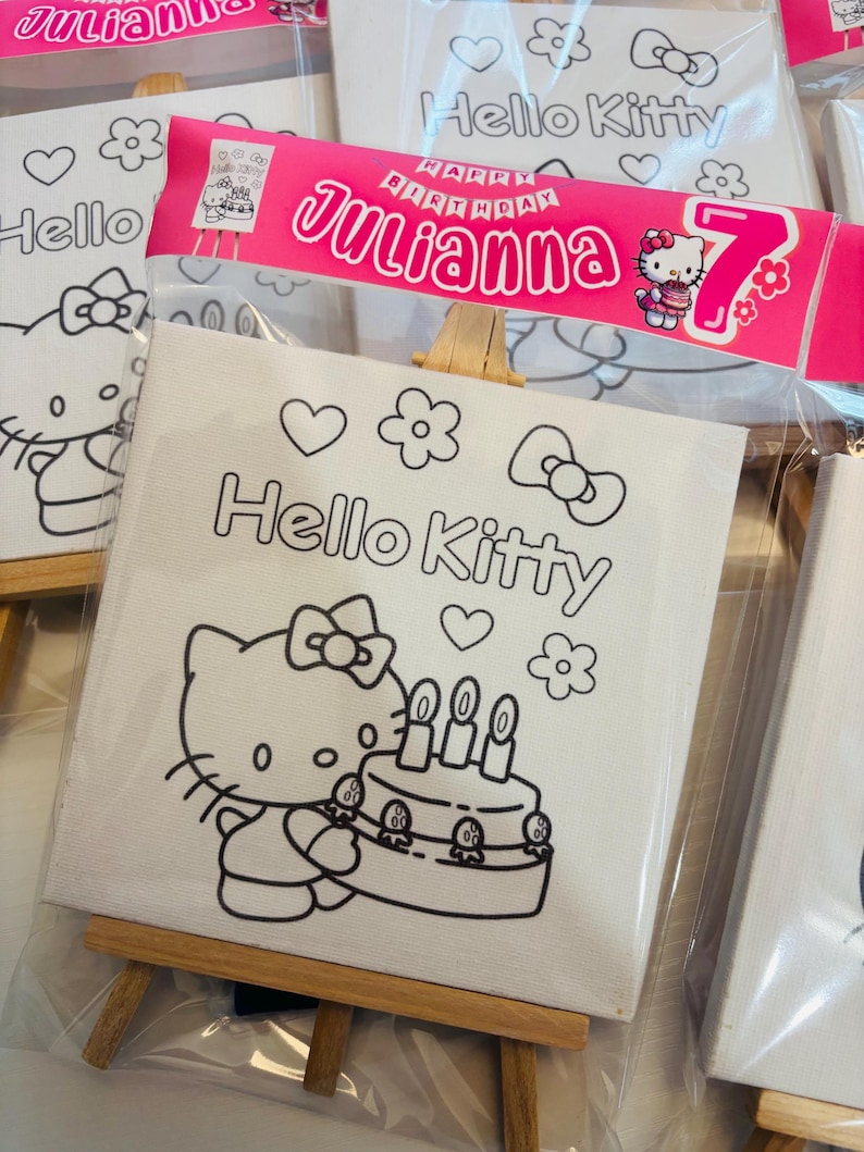 May include: White canvas art featuring a Hello Kitty design, packaged in clear plastic. The design includes Hello Kitty with a birthday cake, hearts, and flowers. A pink banner reads "Happy Birthday Julianna 7".