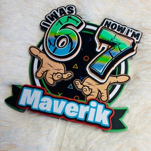 May include: A colorful birthday cake topper with the text "I WAS 6 NOW I'M 7" and the name "Maverik". The design features a black background with green, blue, and white accents, and cartoon hands.