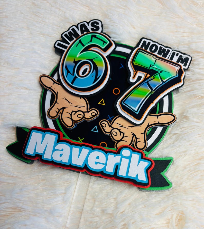 May include: A colorful birthday cake topper with the text "I WAS 6 NOW I'M 7" and the name "Maverik". The design features a black background with green, blue, and white accents, and cartoon hands.