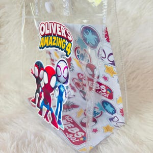May include: Clear plastic tote bag featuring the text "Oliver's Amazing 4" and Spider-Man characters. The bag has a white interior with Spider-Man themed graphics and clear handles.