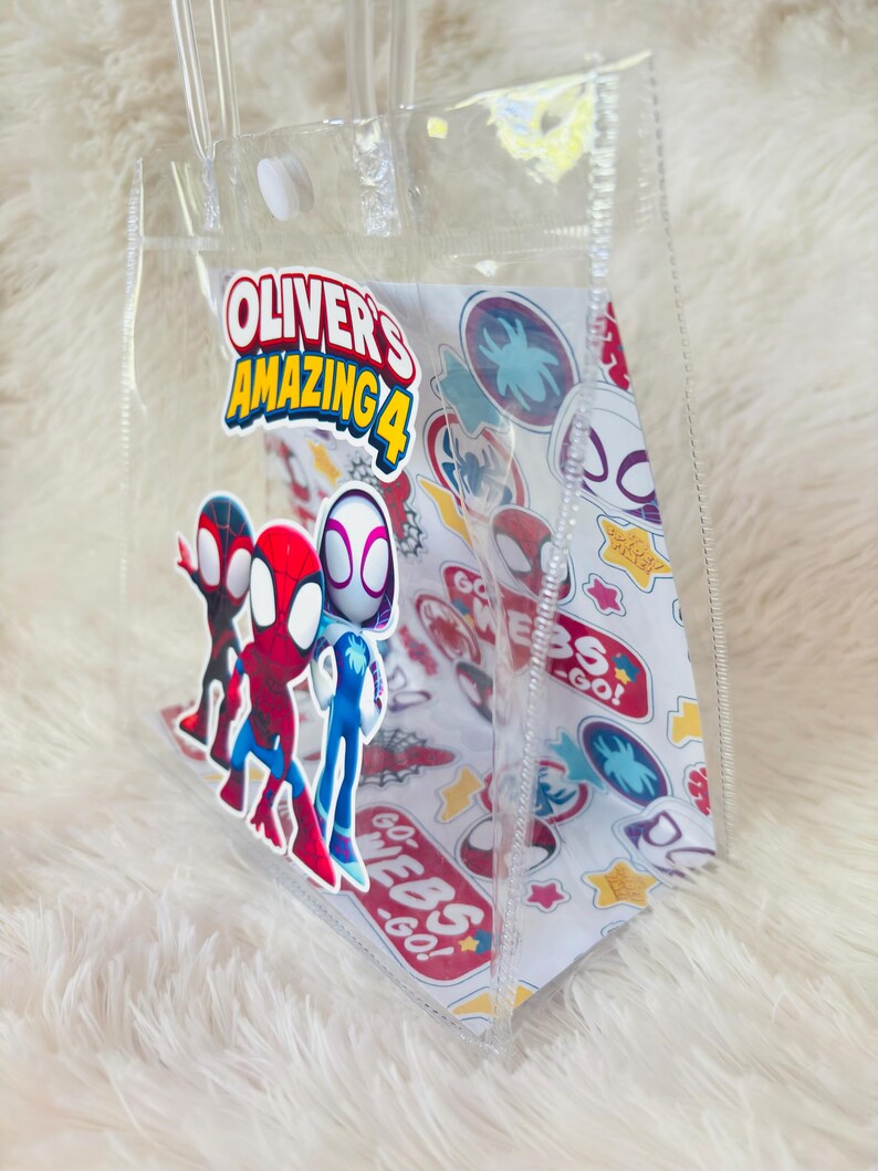 May include: Clear plastic tote bag featuring the text "Oliver's Amazing 4" and Spider-Man characters. The bag has a white interior with Spider-Man themed graphics and clear handles.