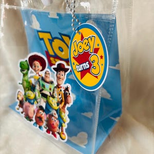 May include: Clear plastic gift bag featuring characters from the movie "Toy Story" against a blue sky background. A round tag with the text "Joey turns 3" is attached. The bag has a white snap closure and clear handles.