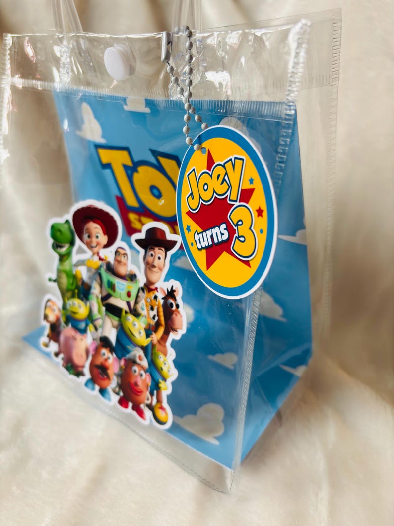 May include: Clear plastic gift bag featuring characters from the movie "Toy Story" against a blue sky background. A round tag with the text "Joey turns 3" is attached. The bag has a white snap closure and clear handles.