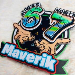 May include: A layered birthday cake topper with the text "I WAS 6 NOW I'M 7" in a graffiti style. The name "Maverik" is also displayed. The design includes hands, geometric shapes, and a black, green, and white color scheme.