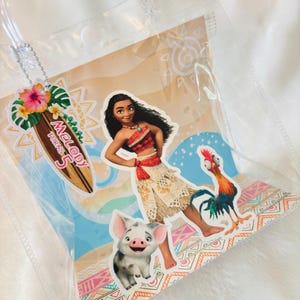 May include: A clear plastic gift bag featuring a Moana-themed graphic with Moana, Pua the pig, and Heihei the rooster. A surfboard-shaped tag attached to the clear handle displays the text Melody Turns 5 with a hibiscus flower and palm leaves.
