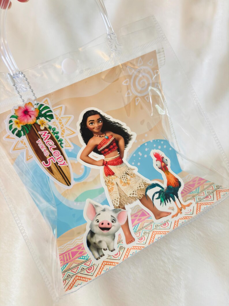 May include: A clear plastic gift bag featuring a Moana-themed graphic with Moana, Pua the pig, and Heihei the rooster. A surfboard-shaped tag attached to the clear handle displays the text Melody Turns 5 with a hibiscus flower and palm leaves.