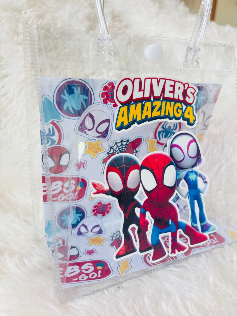 May include: Clear plastic tote bag with a white insert featuring Spider-Man characters and the text "OLIVER'S AMAZING 4". The bag has clear handles and a white snap closure. Colorful stickers of Spider-Man characters are also visible.