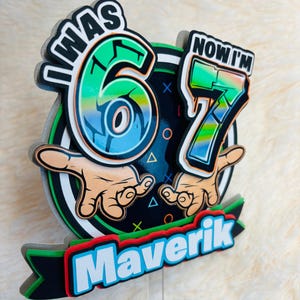 May include: A colorful birthday decoration with the text "I WAS 6 NOW I'M 7" and the name "Maverik". The design features a black background with green, blue, and red accents, and cartoon hands.