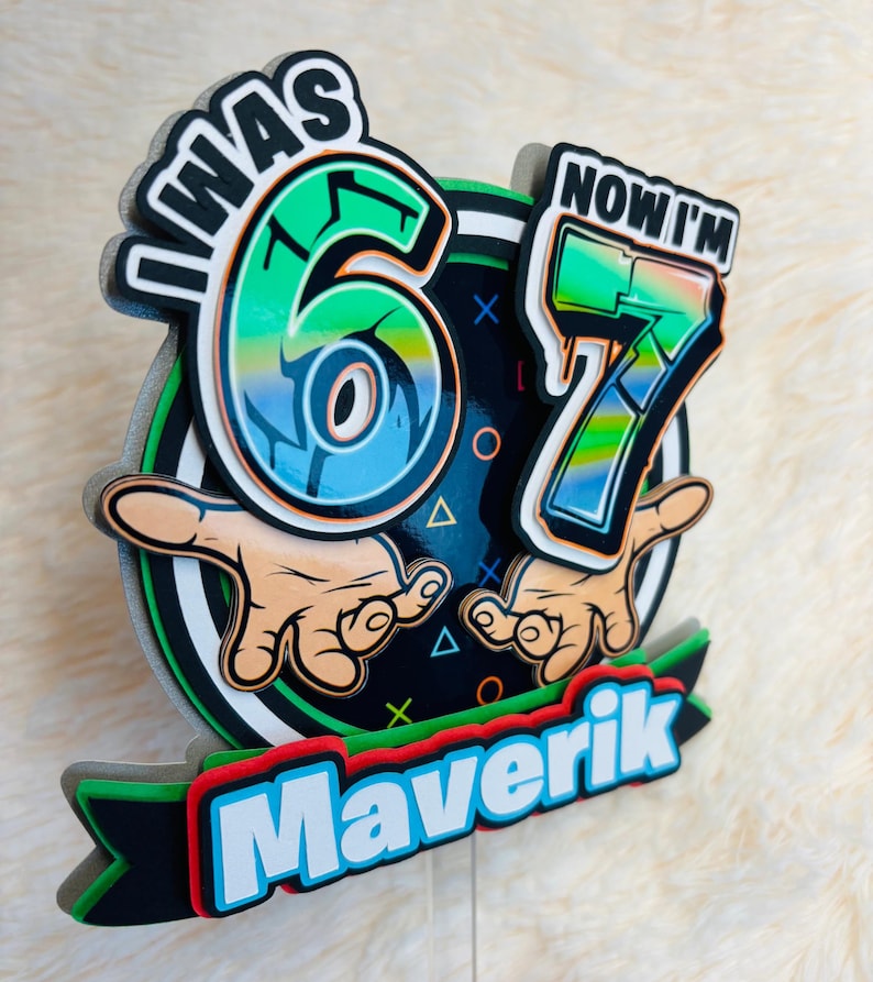 May include: A colorful birthday decoration with the text "I WAS 6 NOW I'M 7" and the name "Maverik". The design features a black background with green, blue, and red accents, and cartoon hands.