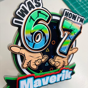 May include: A colorful birthday cake topper with the text "I WAS 6 NOW I'M 7" and the name "Maverik". The design features the numbers 6 and 7 in a vibrant green and blue gradient, with cartoon hands.