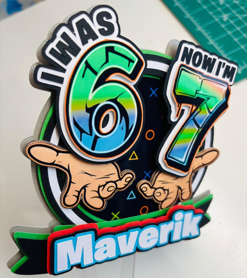 May include: A colorful birthday cake topper with the text "I WAS 6 NOW I'M 7" and the name "Maverik". The design features the numbers 6 and 7 in a vibrant green and blue gradient, with cartoon hands.