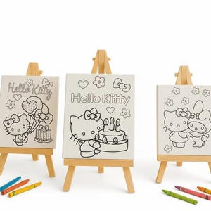 May include: Three small wooden easels each hold a canvas featuring a Hello Kitty design. The canvases are black and white line drawings of Hello Kitty with balloons, a cake, and flowers. Crayons in blue, orange, yellow, green, and red are at the bottom.