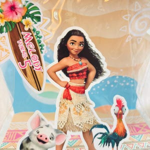 May include: A clear plastic gift bag featuring a Moana character cutout, a piglet, and a rooster. A wooden surfboard-shaped tag attached with a white beaded chain displays the text Melody Turns 5, decorated with a pink hibiscus and green leaves.
