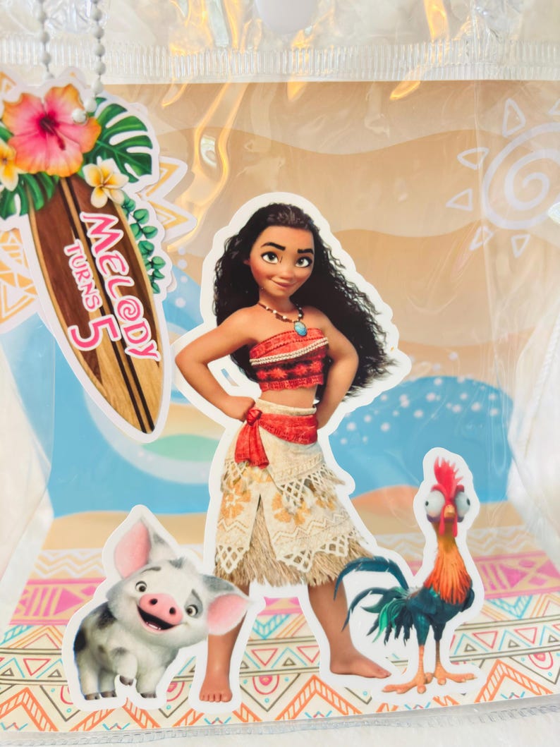 May include: A clear plastic gift bag featuring a Moana character cutout, a piglet, and a rooster. A wooden surfboard-shaped tag attached with a white beaded chain displays the text Melody Turns 5, decorated with a pink hibiscus and green leaves.