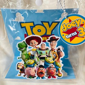 May include: A clear plastic bag containing a blue card with the word "TOY" in yellow letters and various Toy Story characters. A yellow tag with the text "Joey turns 3" is attached.