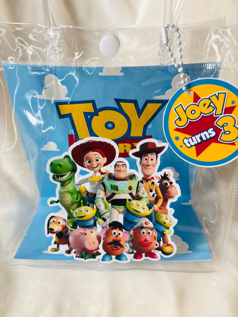 May include: A clear plastic bag containing a blue card with the word "TOY" in yellow letters and various Toy Story characters. A yellow tag with the text "Joey turns 3" is attached.
