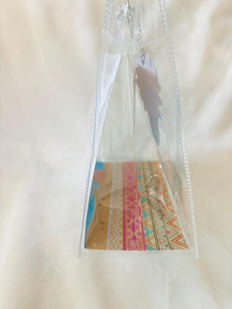 May include: A clear, triangular plastic pouch featuring a colorful base with horizontal bands of geometric patterns in shades of pink, orange, teal, and white. A white, feather-shaped tag hangs from the top zipper closure of the transparent bag.