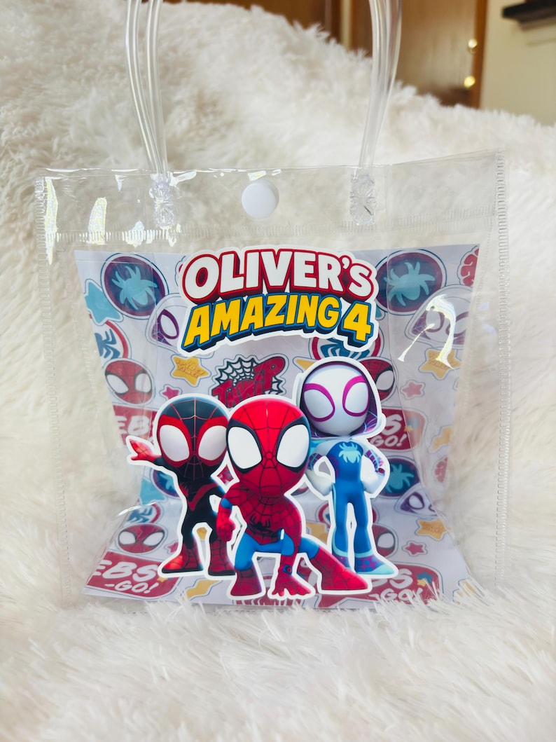 May include: Clear plastic gift bag with a white paper insert featuring the text "Oliver's Amazing 4" and cartoon images of Spider-Man characters. The bag has clear plastic handles and a white button closure.