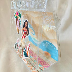 May include: A clear plastic tote bag featuring a Moana-themed graphic insert with a beach scene, a character illustration, and a surfboard-shaped tag that reads Melody Turns 5. The bag has transparent handles and a white snap closure at the top.