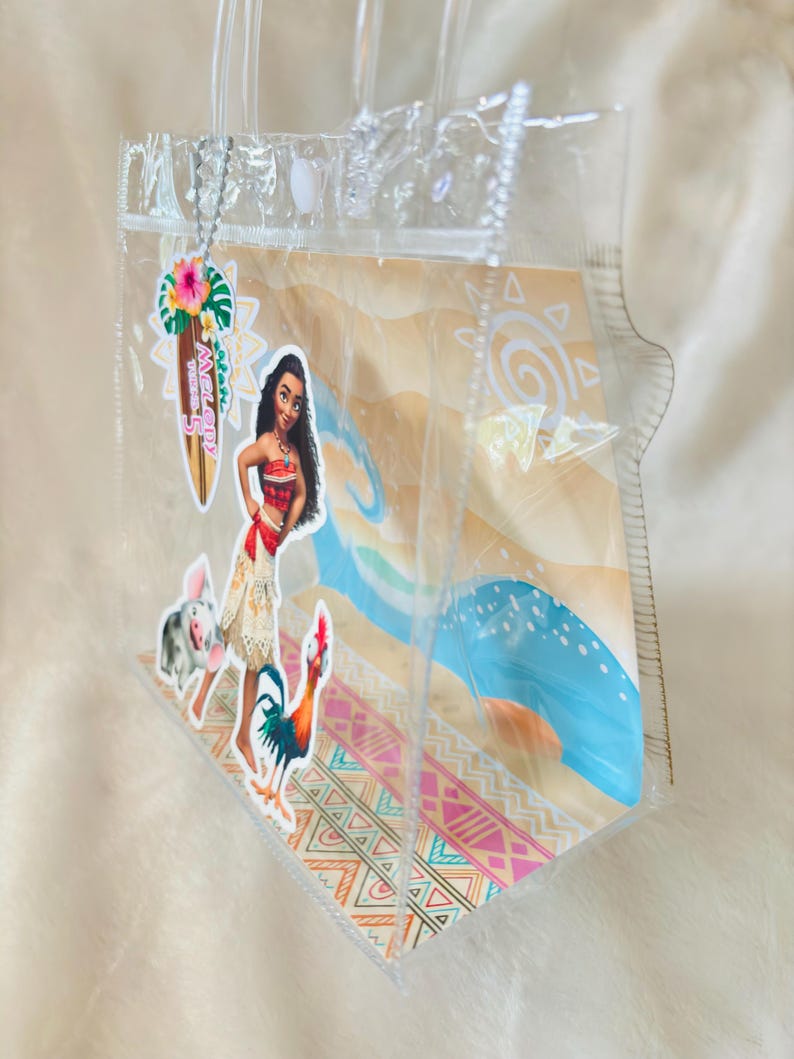 May include: A clear plastic tote bag featuring a Moana-themed graphic insert with a beach scene, a character illustration, and a surfboard-shaped tag that reads Melody Turns 5. The bag has transparent handles and a white snap closure at the top.