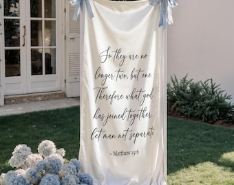 May include: A cream-colored wedding banner with light blue ribbon accents. The banner displays a quote from Matthew 19:6 in elegant black script, along with the names "Kinsey & Sam" and the date "09.13.2026". Decorative blue and white floral arrangements are at the base.