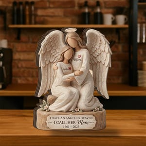 May include: A wooden angel figurine depicting two figures embracing, with large feathered wings. The angel on the right has a halo and the word "Mom" with a heart. The base reads "I HAVE AN ANGEL IN HEAVEN I CALL HER Mom 1961-2025".