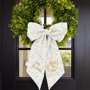 Może przedstawiać: Biała kwiatowa kokarda z napisem "Save The Date" i dwoma splecionymi sercami. Kokarda jest przymocowana do zielonego wieńca na czarnych drzwiach. Na kokardzie wydrukowana jest data 01.02.26.