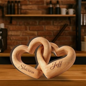 May include: Two wooden heart-shaped sculptures interlocked, with the names "Sharon" and "Jeff" engraved on each. The wood has a light brown color. The background features a brick wall and wooden shelves.