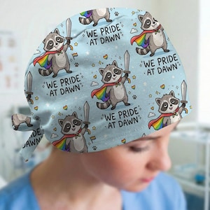 May include: A light blue scrub cap featuring a repeating pattern of cartoon raccoons wearing rainbow capes and holding swords. The text "WE PRIDE AT DAWN" is printed in black lettering alongside small rainbow hearts, stars, and paw prints.