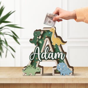 May include: A wooden letter "A" shaped piggy bank with a green interior and dinosaur cutouts. The name "Adam" is written on the front. A hand is inserting a $100 bill into the slot at the top. The dinosaurs are green, yellow, and blue.