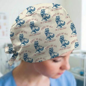 May include: A cream-colored surgical scrub cap featuring a repeating pattern of dancing raccoons in blue and white. The text "FIRST OF ALL I'M A DELIGHT" is printed around the raccoons. The cap has a tie closure.