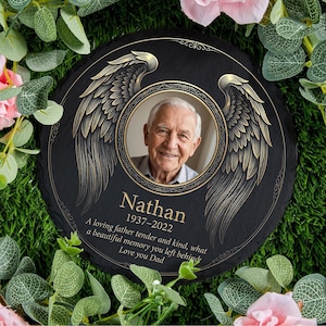 Op de afbeelding: Een zwarte gedenksteen met een foto van een glimlachende oudere man, omringd door goudkleurige engelen vleugels. De steen bevat de naam "Nathan", data 1937-2022 en een liefdevolle boodschap. Omringd door groen en roze bloemen.