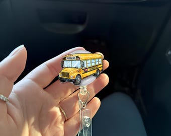 Puede incluir: Un carrete de insignia de autobús escolar amarillo con un clip de plástico transparente. El gráfico del autobús escolar tiene las palabras "School Bus" en la parte superior y lateral. El carrete es redondo y blanco.