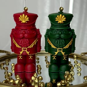 May include: Two decorative candles shaped like toy soldiers. One is red, the other green, both with gold accents. They are on a mirrored tray with gold bells scattered around. The candles have a detailed design, including hats and belts.