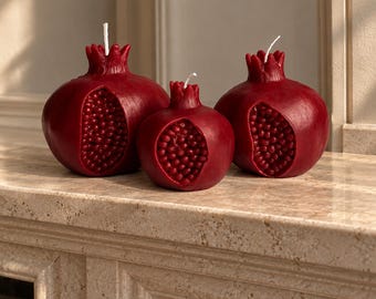 May include: Three deep red pomegranate-shaped candles of varying sizes. Each candle features a textured exterior with a realistic open section revealing individual seeds and a small white wick protruding from the top crown of the fruit.