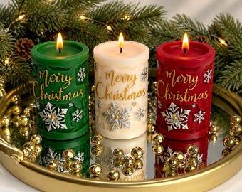 May include: Three cylindrical pillar candles in green, cream, and red, each featuring a lit wick, gold embossed Merry Christmas text, and silver snowflake accents. The candles are arranged on a round gold tray with small gold decorative spheres.